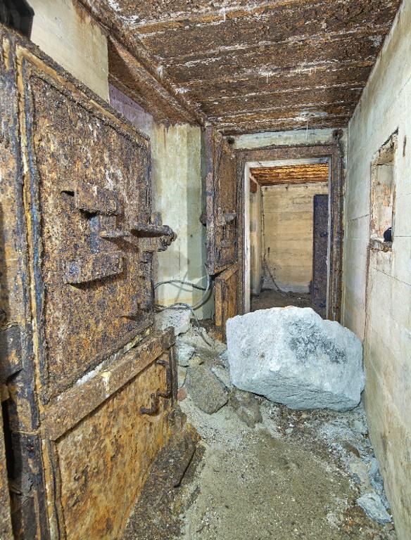 Vue du débouché du couloir de l'entrée sud : il est encombré de gravas. Au premier plan à gauche, porte blindée à deux vantaux du sas de la salle de garde. Au fond, la porte blindée du sas de la caponnière et du magasin à munitions
