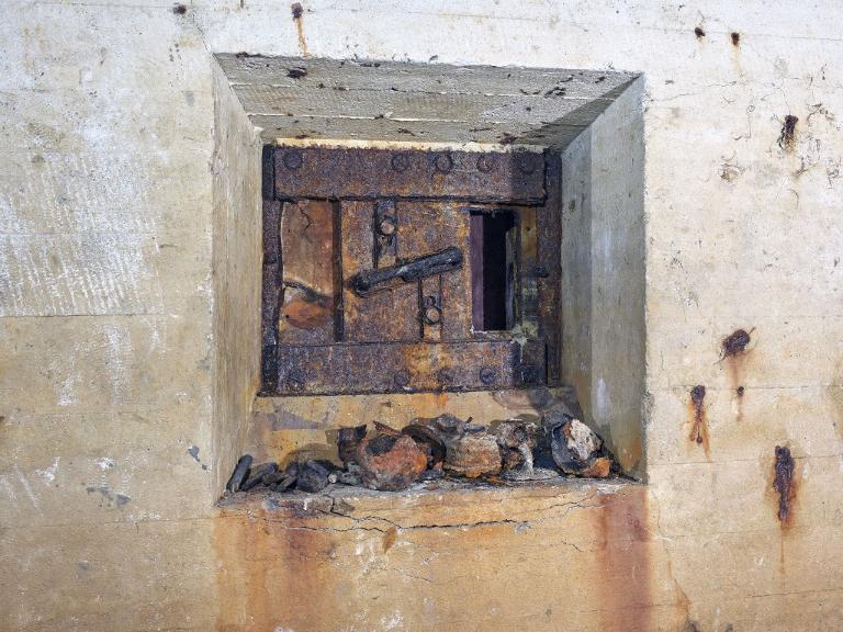 Vue de détail de l'ouverture de tir blindée de l'entrée ouest. Sur le rebord se trouvent regroupés différents artéfacts : brosses en bois, cartouches de masque à gaz...