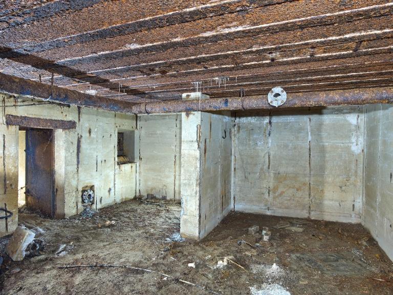 Vue de la salle de garde : elle est partiellement divisée par un mur de refend. A gauche, porte étanche d'accès et bouche de surpression à clapet. Au plafond, tuyauterie du système de ventilation
